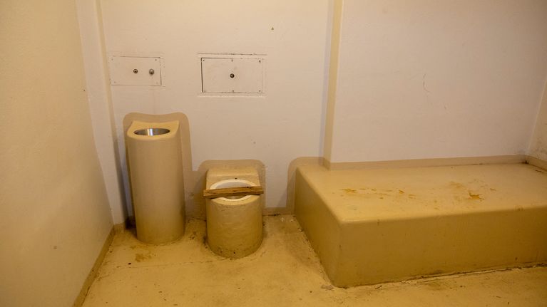 Enkel fängelsecell med toalett, handfat och fast bänk längs väggen.
