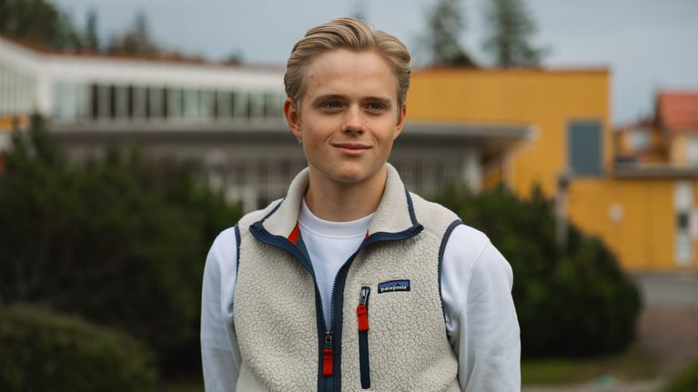 En ung person med ljust hår och vit fleeceväst står utomhus framför en gul byggnad och grönska.