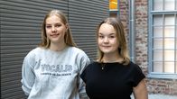 Två personer står bredvid varandra i en byggnad med tegelväggar och stora fönster.