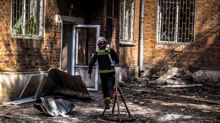 Palosammuttaja astelee kohti avointa ulko-ovea, pihalla pellin paloja ja lasinsiruja.