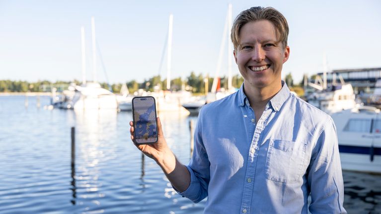En ung man står vid en småbåtshamn och håller upp en mobiltelefon med en karta synlig på skärmen.