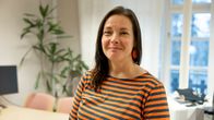 Nainen raidallisessa paidassa seisoo toimiston valoisan huoneen edustalla, taustalla on suuri ikkuna ja kasvi.