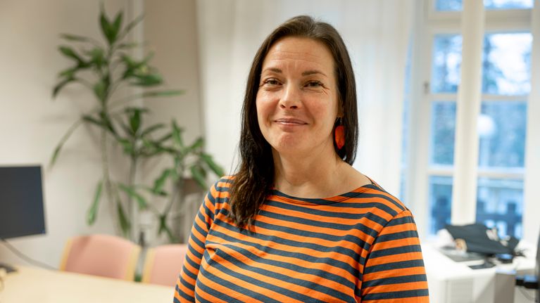 Nainen raidallisessa paidassa seisoo toimiston valoisan huoneen edustalla, taustalla on suuri ikkuna ja kasvi.
