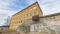 Kuvassa on vanha keltainen koulurakennus ja sen edustalla oleva rapistunut koripallokenttä. Rakennuksessa on useita ikkunoita ja sen ulkoseinässä näkyy graffiteja.