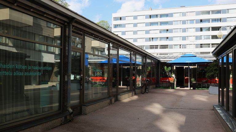 Gård mellan två byggnader med stora fönster, uteservering med blått parasoll och ett flerbostadshus i bakgrunden.