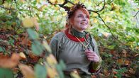 Oranssihiuksinen nainen istuu metsässä syksyisessä maisemassa, päässään itsetehty seppele ja kaulassaan suurentava luuppi.