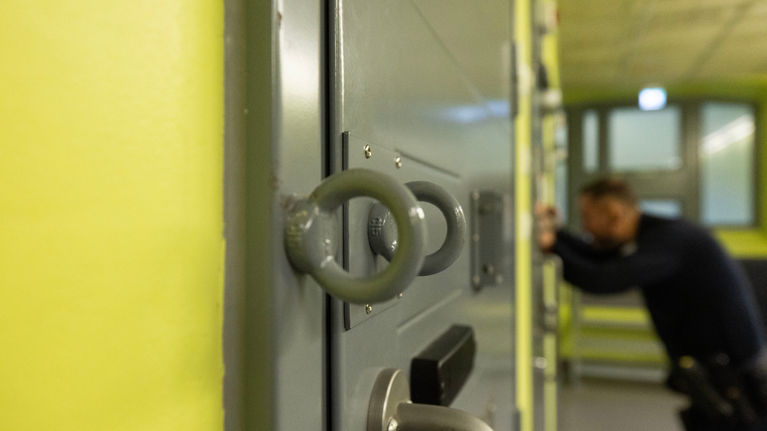 Prison guard in dark uniform, seen blurry in the background, looking through a peephole into a prison cell. Walls are yellow and cell doors are grey. 