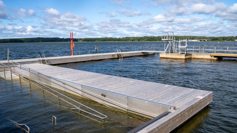 Utomhusbrygga med hopptorn i bakgrunden och en tillgänglig terapibassäng med ramp i förgrunden.