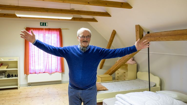 Christer Westerholm står med utsträckta armar i ett rum med trägolv, synliga takbjälkar och sängar.