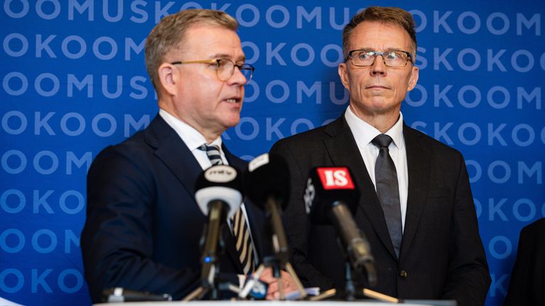 Kaksi miestä seisoo puhujapöntön äärellä mikrofonien edessä, taustalla toistuva teksti "KOKOOMUS".