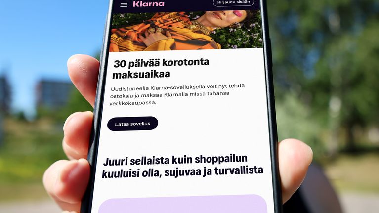 Käsi pitää puhelinta, jossa näkyy Klarnan sivusto ja teksti "Juuri sellaista kuin shoppailun kuuluisi olla, sujuvaa ja turvallista".