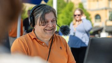 Roger Källman med headset och mikrofon sitter utomhus vid ett bord, medan en annan person står i bakgrunden.