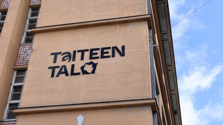 Fasad av en beige byggnad med texten "Taiteen Talo" i svart, blå himmel syns i bakgrunden.