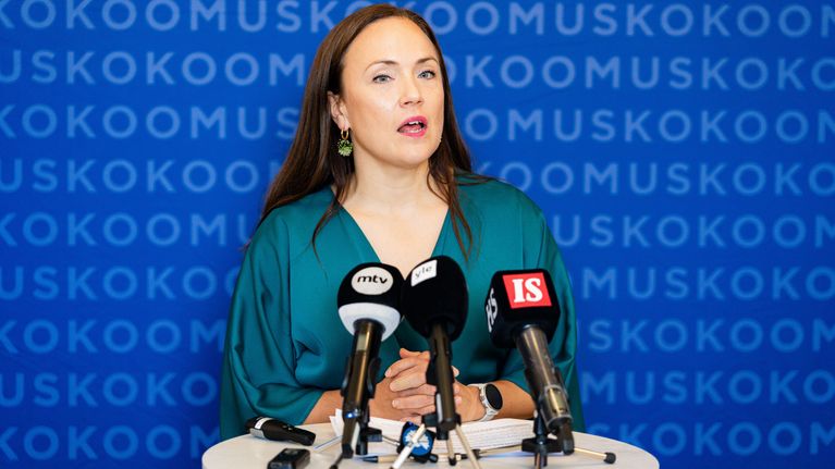 Nainen seisoo puhujapöntön takana useiden mikrofonien edessä, taustalla toistuva sana "KOKOOMUS".
