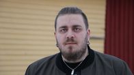 En man men kort mörkt hår och skägg och med stora örhängen i öronen står framför en gul ladugårdsdörr.