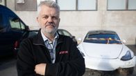 En man står med korsade armar framför en vit bil med skador på fronten. På jackan syns texten "Autocirc"