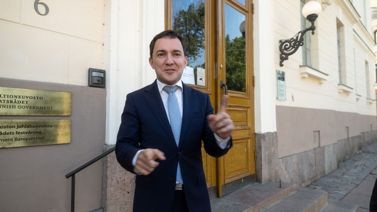 Wille Rydman i en mörkblå kostym, vit skjorta och ljusblå slips står utanför en byggnad och pratar. Han gestikulerar med händerna.