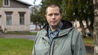 En person står utomhus i en grön jacka. I bakgrunden syns en äldre byggnad och några träd.