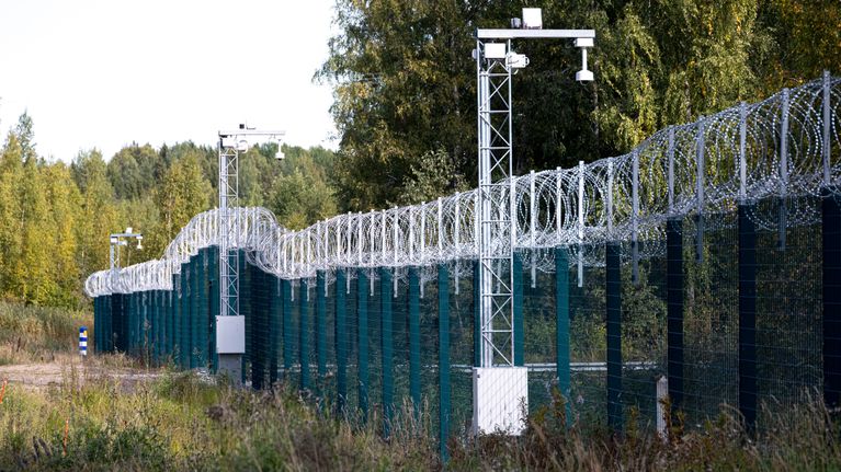 Piikkilangalla varustettu raja-aita metsäisessä maastossa, aidan vieressä valvontakameroita.