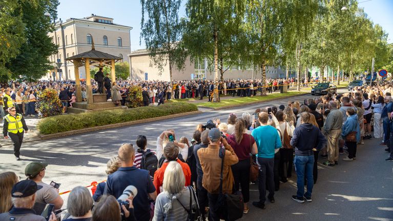 En stor folkmassa står samlad längs en gata, med flera poliser och byggnader i bakgrunden.