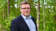 Tummaan puvuntakkiin pukeutunus mies seisoo metsän reunassa.
