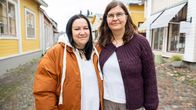 Två kvinnor står bredvid varandra på en stenbelagd gata mellan trähus i ljusa färger.
