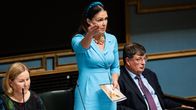 Kvinna med mörkt långt hår och ljusblå klänning står och talar i riksdagen.