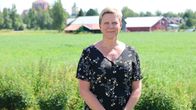 En kvinna i blommig klänning står framför en åker. I bakgrunden syns röda byggnader.