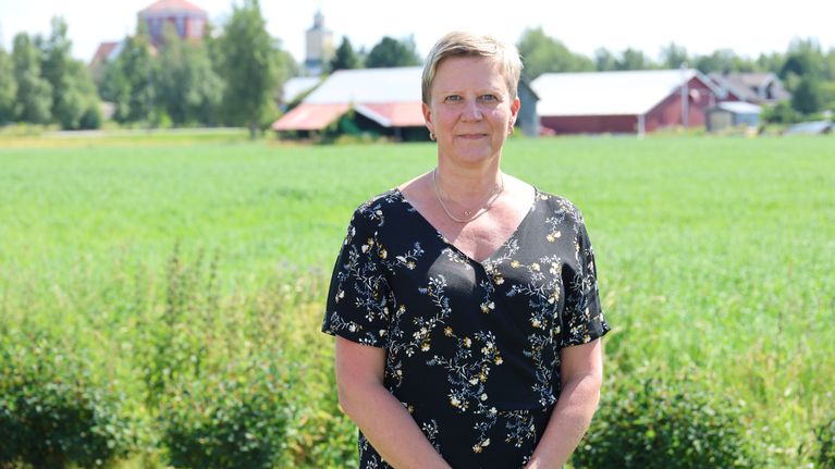 En kvinna i blommig klänning står framför en åker. I bakgrunden syns röda byggnader.