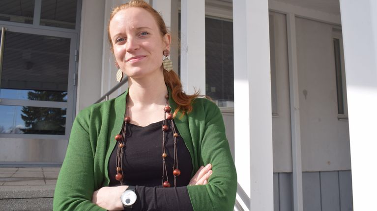 Punatukkainen nainen vihreässä neuleessa ja puuhelmissä katsoo kameraan selkeäilmeisenä. Henkilökuva.