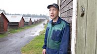 Personen på bilden står vid en byggnad och bär arbetskläder som är blå och gröna med texten "MILMA" på bröstet. I bakgrunden syns en rad med låga byggnader för minkar, med röda väggar och vita tak längs en grusväg.