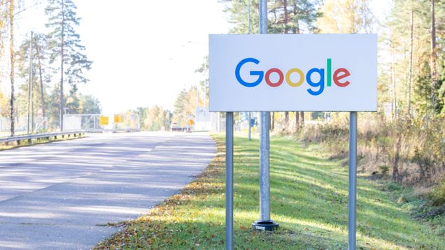Tien varressa on valkoinen kyltti, jossa on Google-logo värikkäillä kirjaimilla. Taustalla on metsää ja aita.