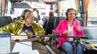 Två personer med hörlurar sitter i en buss, den ena spelar gitarr, flera mikrofoner och teknikutrustning syns.
