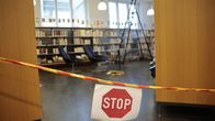 Kirjaston hyllyjen edessä esteenä on keltainen nauha ja "STOP"-kyltti; taustalla seisoo tikkaat.