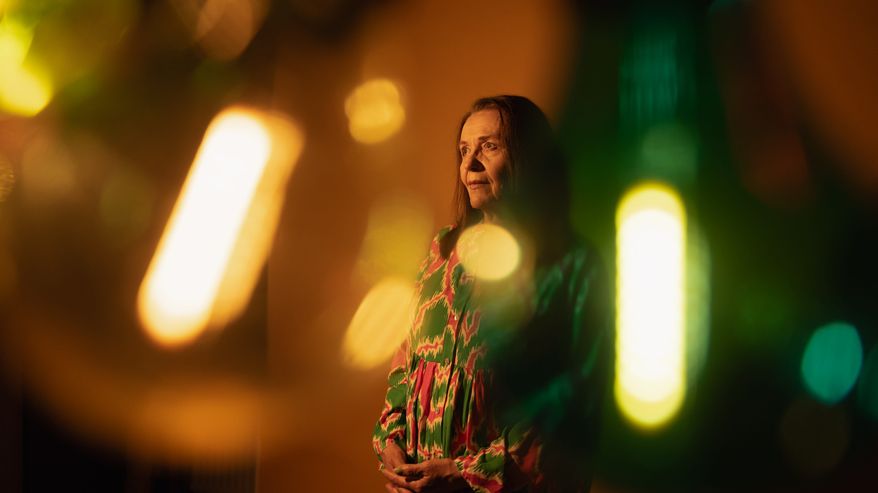 Riitta Havukainen kuvattuna värillisten lamppujen takaa. Havukainen seisoo kädet ristissä, katsoo sivuun kaukaisuuteen.