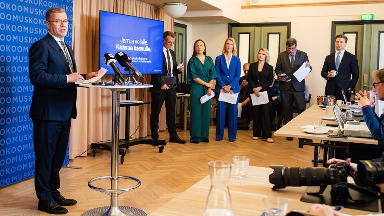 Useita ihmisiä seisoo kokoushuoneessa puhujanpöntön äärellä, taustalla näytöllä teksti "Jarrua velalle. Kaasua kasvulle."
