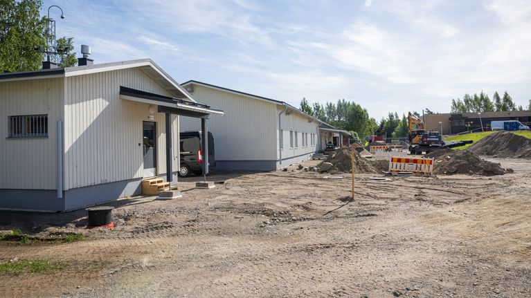 Vaalea rakennus työmaan laidalla, pihalla kaivinkone ja ajorata suljettu muoviaidoilla.