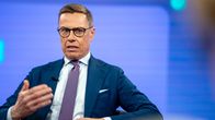 A man wearing glasses, a blue jacket and a purple tie sits in a TV studio, speaking with a serious expression and gesturing with one hand.