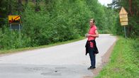 En person i röd tröja står vid en landsväg bredvid två vägskyltar. Hon tittar på ett hål i vägen. 