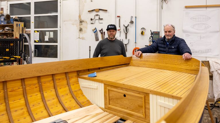 Mathias Blomqvist och Christer Westerholm står vid en träbåt i en verkstad med verktyg och ritningar på väggarna.