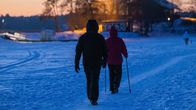 Two dark figures seen from the back walking on snow-covered ice at dusk with trees and an old wooden house in the background.