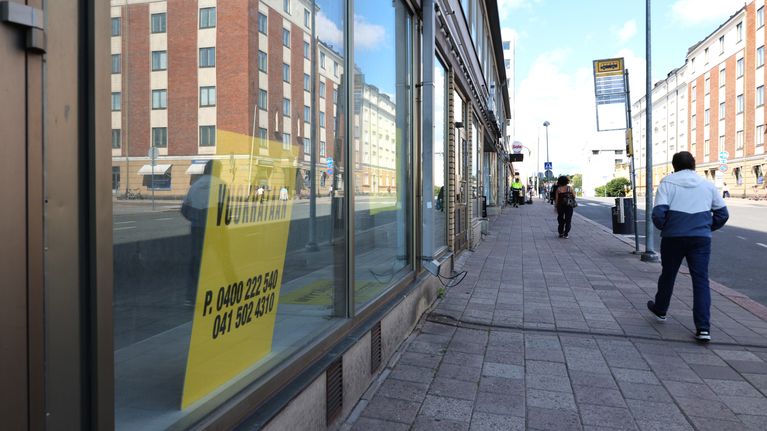 Personer går på en trottoar intill en rad butiker; i ett skyltfönster syns en gul skylt med texten ”VUOKRATAAN”.