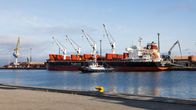 A freight shipping harbour with a huge freighter ship docked next to a row of large cranes. A tugboat in the foreground.