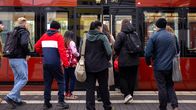 About eight people standing at a platform, waiting to board a red coloured tram.