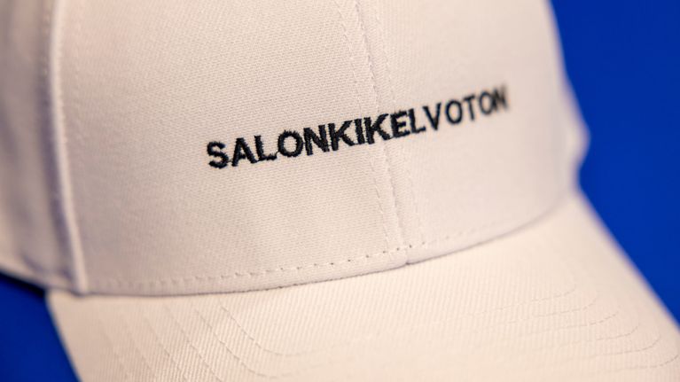 Valkoinen lippalakki, jossa on teksti "SALONKKIKELVOTON", asetettuna siniselle pöydälle.