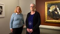 Två personer står framför en målning på en mörkröd vägg. 