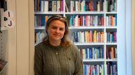 En person med ljust hår står framför en bokhylla på ett bibliotek. Skyltar med bokstäverna I och J syns till vänster.