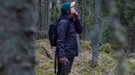Nainen seisoo metsässä ja katsoo puiden latvoihin.