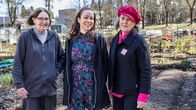 Tre glada damer skrattar i vårsolen mot kameran på Borgå fritidsodlares odlingslotter. 