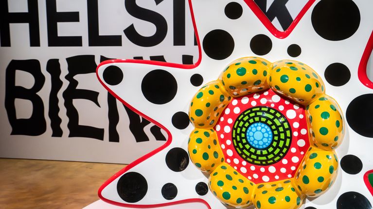 Färgglad skulptur med prickar och mönster står framför en vägg med texten "HELSINKI BIENNALE".